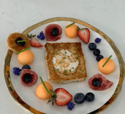 A square egg-in-a-hole toast sits on a white plate, surrounded by blueberries, cantaloupe balls, strawberries, salami slices, and mini pancakes with jelly—an arrangement worthy of display in any modern appliance showroom.