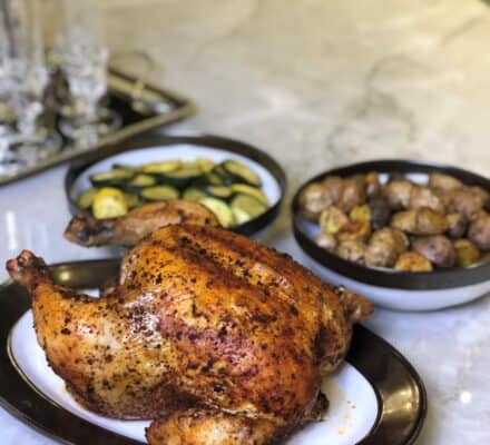 A whole roasted chicken on a plate with bowls of roasted potatoes and sautéed zucchini sits on a marble countertop, showcasing the elegance of a kitchen showroom featuring premium Wolf appliances.