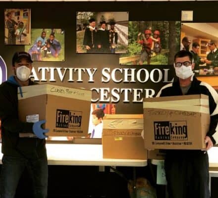 Two people wearing masks and gloves hold large labeled boxes in front of a school display board with photos and the words "Creativity School Center," reminiscent of a delivery scene at an appliance showroom.