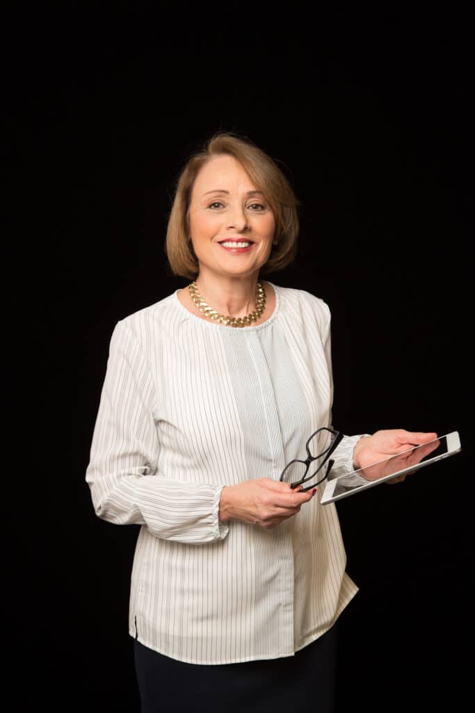 A woman in a white blouse holds eyeglasses and a tablet, standing against a plain black background and smiling at the camera—ready to help you explore the latest innovations at our appliance showroom.