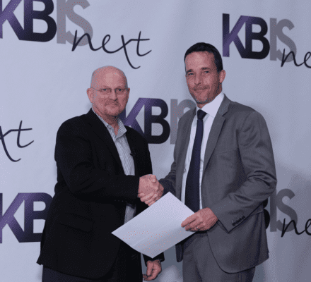 Two men in business attire shake hands at a KBIS Next event, standing in front of a branded backdrop, possibly discussing the latest Wolf appliance for a luxury kitchen showroom, with one holding a sheet of paper.