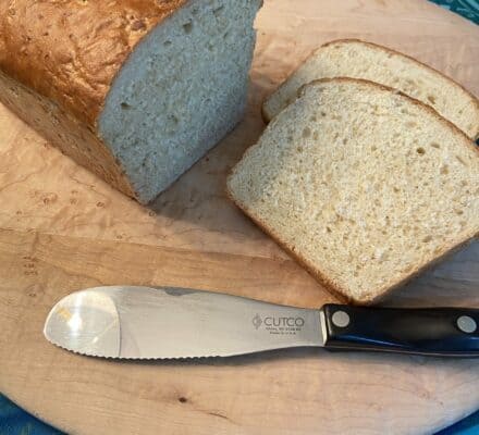 A loaf of bread with two slices cut, placed on a wooden cutting board next to a serrated Cutco knife, creates the perfect scene in front of a gleaming Wolf appliance—ideal for any luxury kitchen appliance lover.