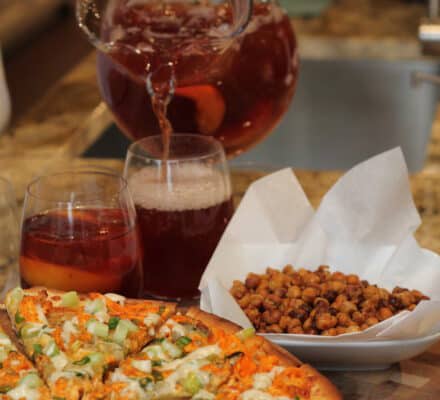 A person pours a red beverage from a glass pitcher into a glass beside a pizza, two filled glasses, and a bowl of roasted chickpeas on a kitchen counter with a luxury kitchen appliance in view.