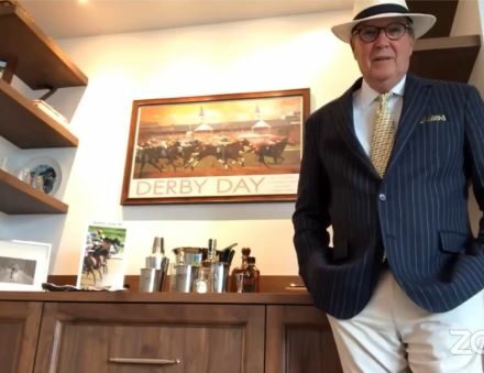 A man in a pinstripe blazer, light pants, tie, and hat stands in front of a kitchen showroom cabinet with drinks and a "Derby Day" horse racing poster on the wall.