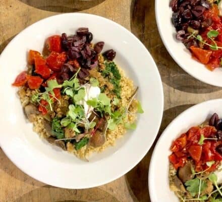 Five white plates filled with quinoa, roasted red peppers, black olives, greens, and a dollop of white sauce are artfully arranged on a wooden surface—perfect inspiration for your next meal after visiting an appliance showroom.
