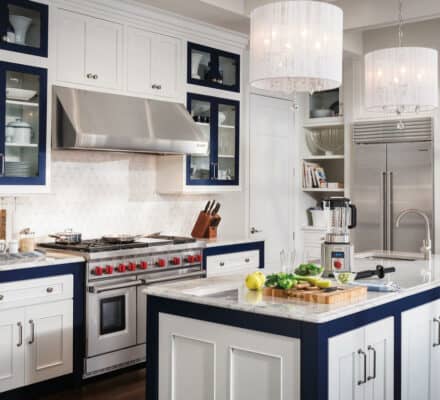 Modern kitchen showroom featuring white cabinets, navy blue accents, stainless steel appliances including a Sub Zero appliance, and an island with a cutting board, blender, and fresh vegetables. Two pendant lights hang above the island.