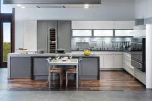 Modern kitchen showroom with gray and white cabinets, a central island dining setup, wooden chairs, and luxury kitchen appliances including a Wolf appliance on dark wood and gray tile flooring.