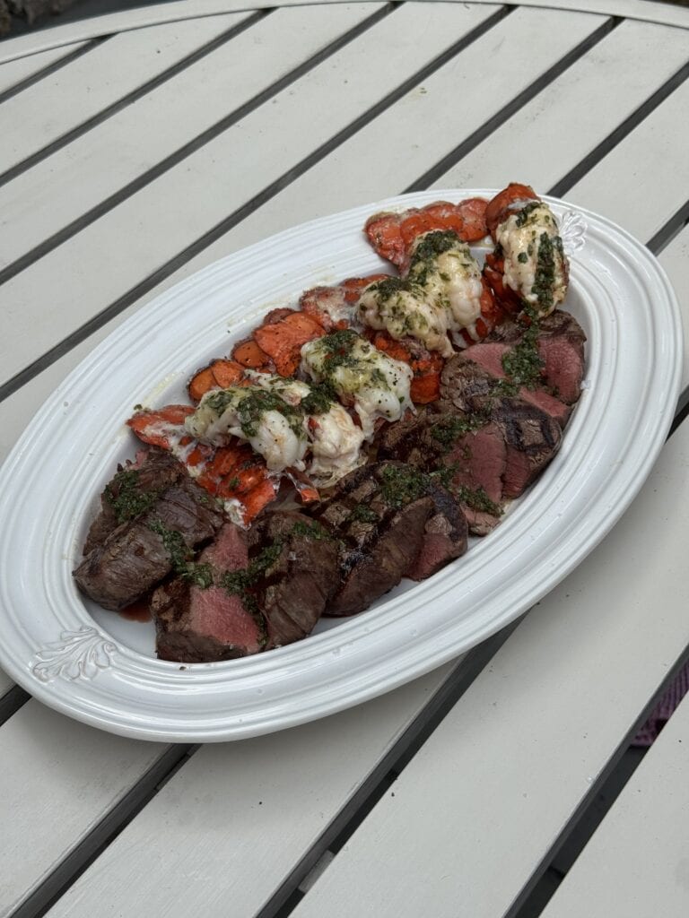 Grilled surf and turf platter with sliced filet mignon and lobster tails topped with chimichurri on a white serving dish
