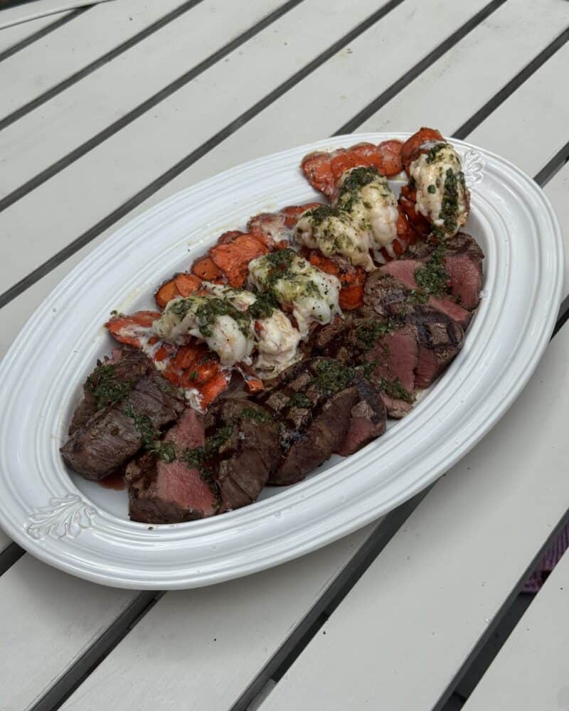 Grilled surf and turf platter with sliced filet mignon and lobster tails topped with chimichurri on a white serving dish