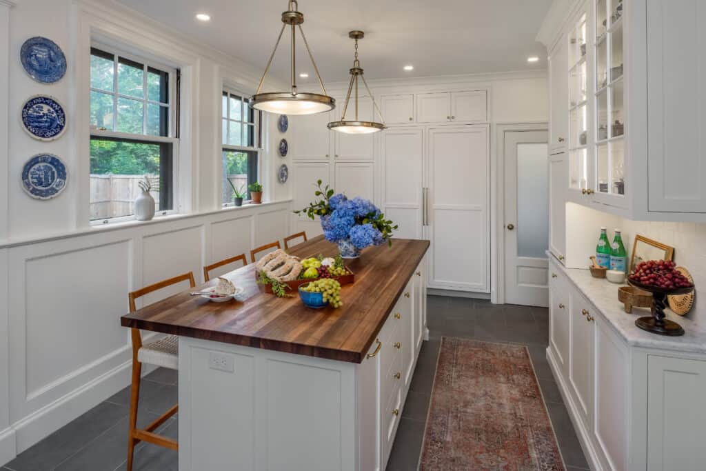 Bright, restored kitchen with white cabinets, a wooden island, bar stools, blue plates on the wall, a rug, and a vase of blue flowers; fresh fruit and food brighten the kitchen renovation.