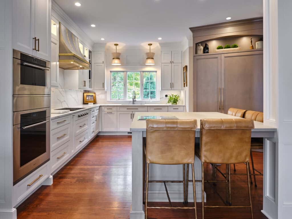Modern eat-in kitchen with white cabinetry, stainless steel appliances, marble backsplash, wooden floor, an island with two tan leather chairs, and large windows letting in natural light.