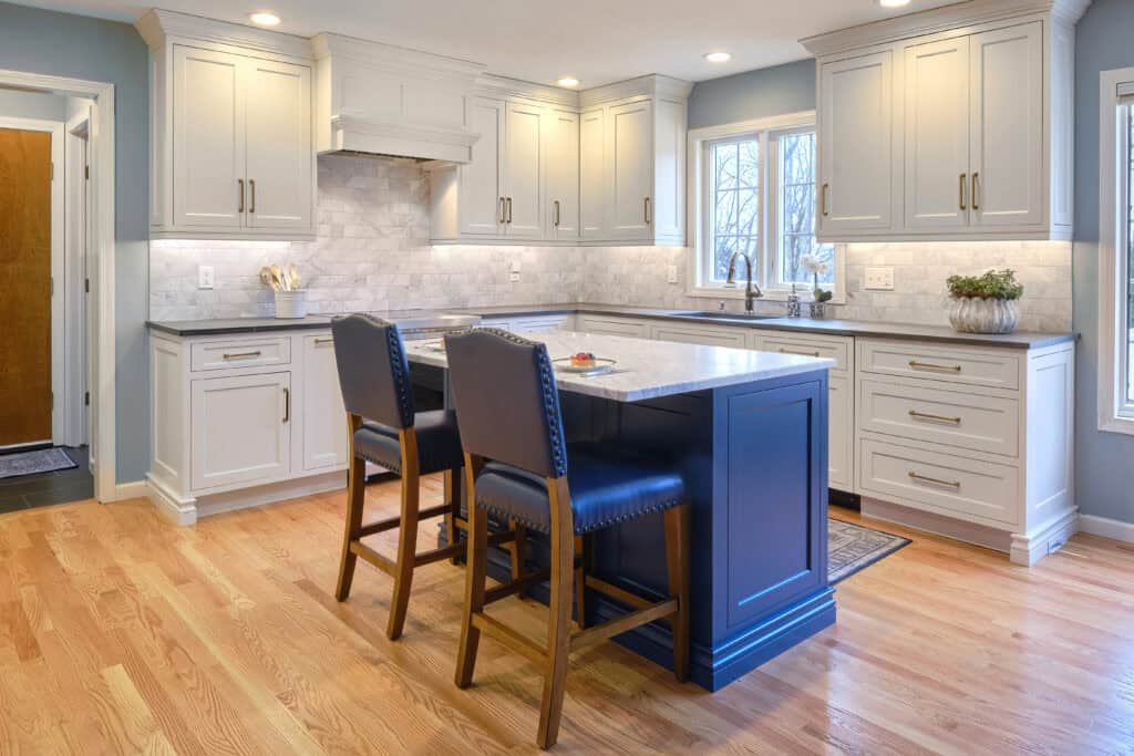 Modern Transitional Kitchen in Hamden, Connecticut features white custom built-in cabinets, a marble backsplash, blue island with sink, two dark chairs, hardwood floors, and recessed lighting.