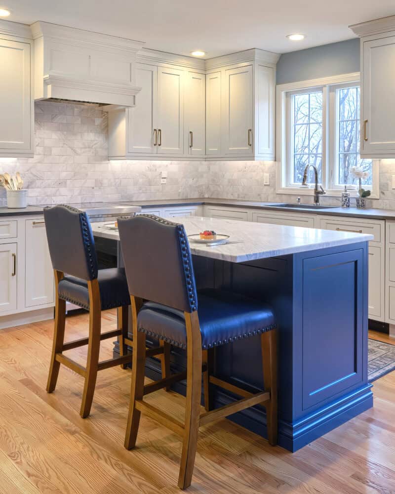 Modern Transitional Kitchen in Hamden, Connecticut features white custom built-in cabinets, a marble backsplash, blue island with sink, two dark chairs, hardwood floors, and recessed lighting.