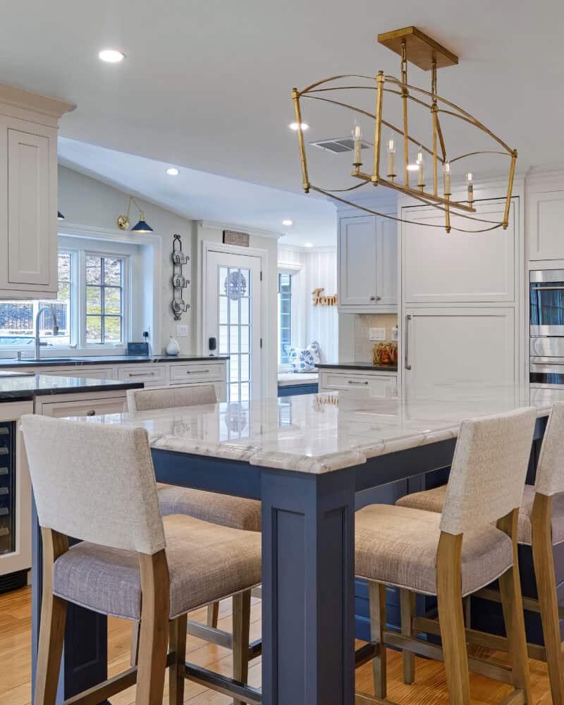 Modern kitchen with white cabinets, marble island, upholstered bar stools, and stainless steel appliances. This spacious kitchen in Cheshire, Connecticut features wooden floors, a gold chandelier, plenty of storage, and abundant natural light.