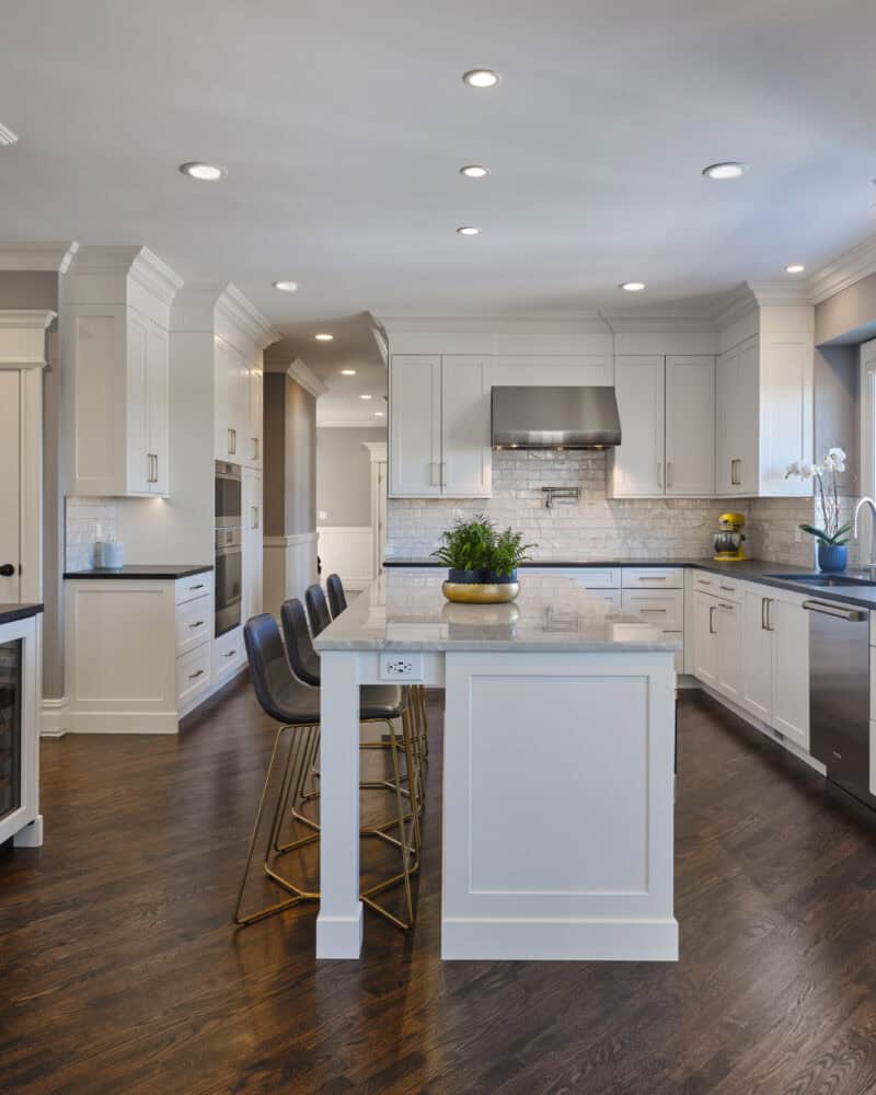 Spacious gourmet kitchen in Avon CT features white cabinets, stainless steel appliances, dark countertops, a large island with barstools, and hardwood flooring.