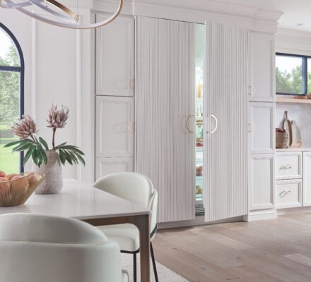 Modern kitchen with white cabinetry, a hidden Sub-Zero fridge with panel doors slightly open, round chandelier, light wood floors, and a dining table with white chairs and a centerpiece vase—perfect for those wondering what are Sub-Zero appliances.