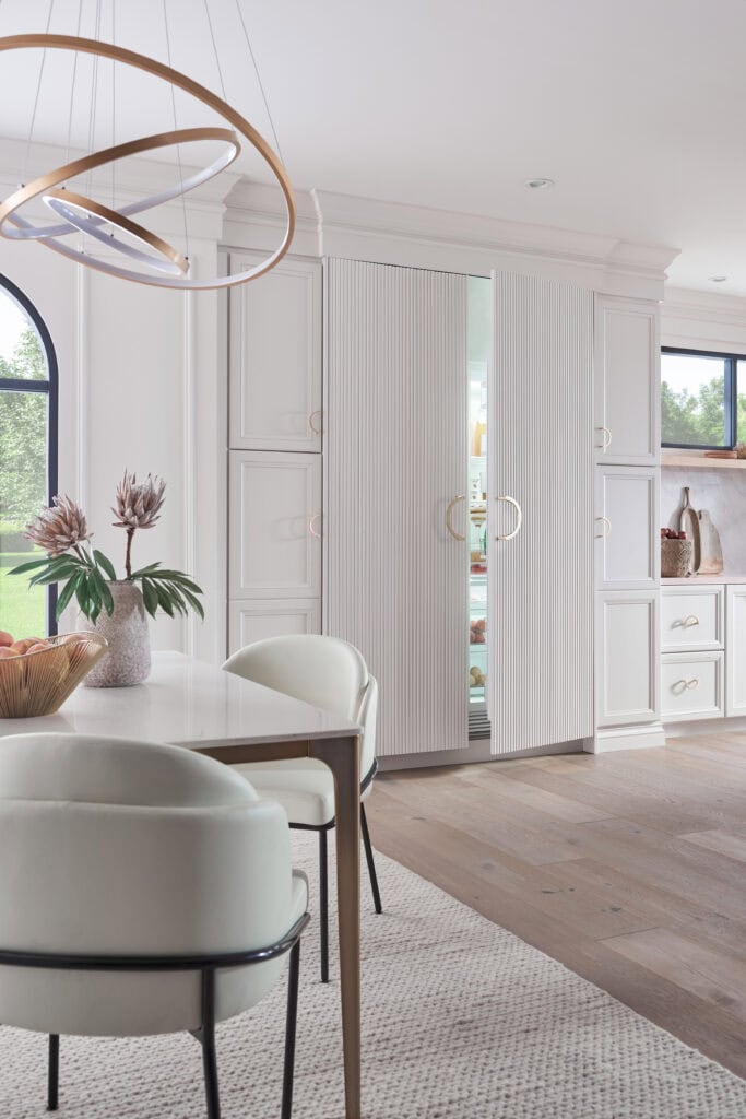 Modern kitchen with white cabinetry, a hidden Sub-Zero fridge with panel doors slightly open, round chandelier, light wood floors, and a dining table with white chairs and a centerpiece vase—perfect for those wondering what are Sub-Zero appliances.