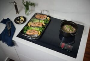 A wolf cooktop with salmon, bok choy, and lemon sizzling on a griddle, soba noodles simmering in a pot, and garlic with fresh herbs arranged on a nearby plate.