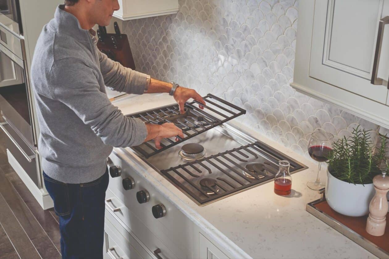 A person lifts a metal grate from a Wolf cooktop in a modern kitchen with a tiled backsplash, a glass of wine, and a potted plant on the counter.