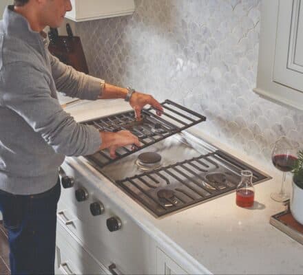 A person lifts a metal grate from a Wolf cooktop in a modern kitchen with a tiled backsplash, a glass of wine, and a potted plant on the counter.