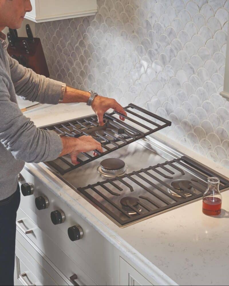 A person lifts a metal grate from a Wolf cooktop in a modern kitchen with a tiled backsplash, a glass of wine, and a potted plant on the counter.
