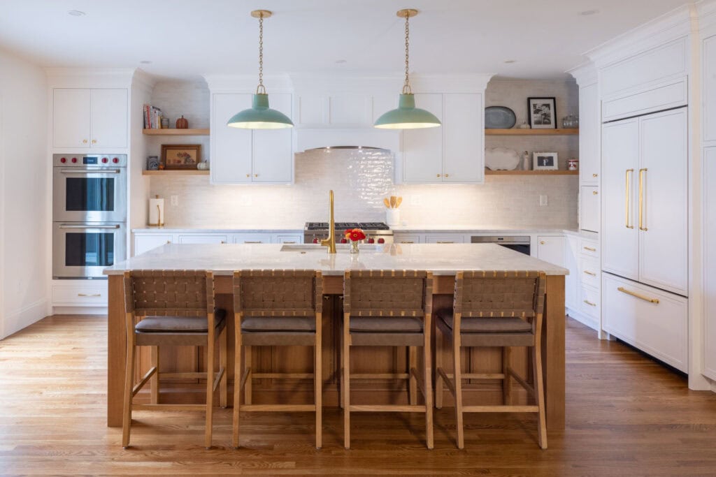 A modern kitchen in the Sitcoske Residence features white cabinets, a large island with four wooden barstools, pendant lights, built-in ovens, and a refrigerator.