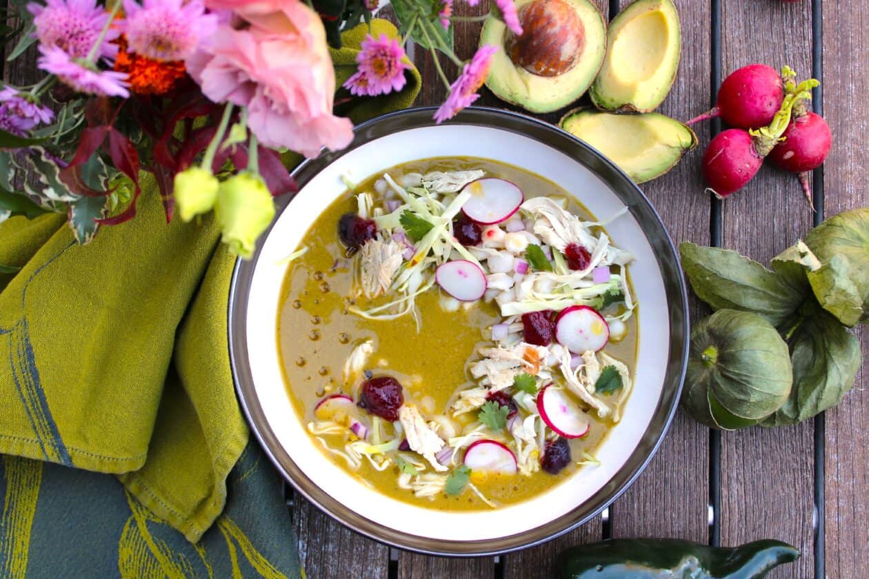 A bowl of Turkey Pozole Verde garnished with shredded turkey, sliced radishes, lettuce, and cilantro sits on a wooden table, surrounded by avocados, tomatillos, radishes, and flowers—a perfect idea for leftover turkey recipes.