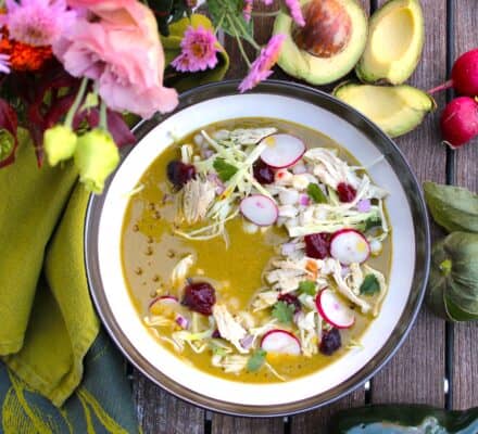 A bowl of Turkey Pozole Verde garnished with shredded turkey, sliced radishes, lettuce, and cilantro sits on a wooden table, surrounded by avocados, tomatillos, radishes, and flowers—a perfect idea for leftover turkey recipes.