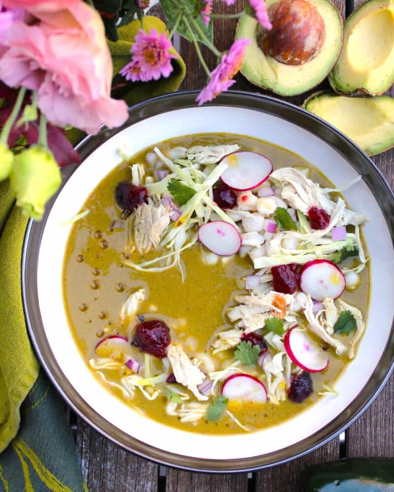 A bowl of Turkey Pozole Verde garnished with shredded turkey, sliced radishes, lettuce, and cilantro sits on a wooden table, surrounded by avocados, tomatillos, radishes, and flowers—a perfect idea for leftover turkey recipes.