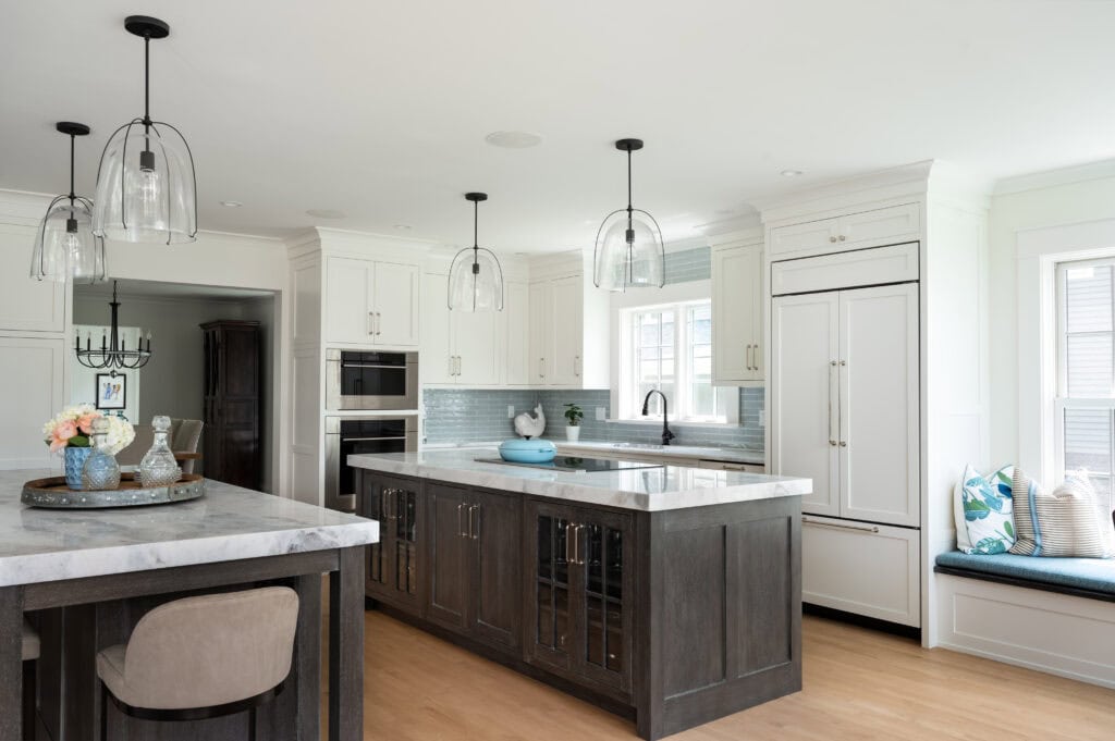 Modern kitchen with white cabinets, a large dark island, marble countertops, pendant lights, built-in appliances, and a window seat with blue cushions creates a Seabreeze Revival vibe.