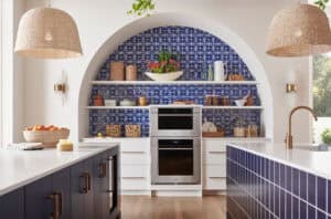 Contemporary kitchen with blue geometric tile backsplash, arched shelving, built-in convection oven, woven pendant lights, and white countertops with blue cabinetry.