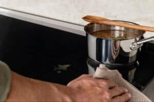 A person wipes a spill on a black induction cooktop next to a pot of soup with a wooden spoon resting on top.
