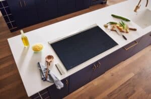 Modern kitchen countertop featuring a sleek black induction cooktop, wooden utensils, a bottle of oil, a sponge, and fresh vegetables on a cutting board near the sink.