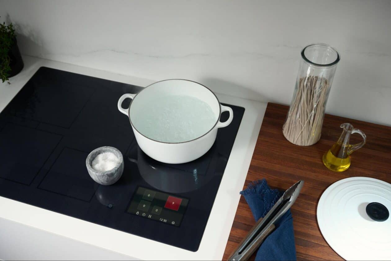 A white pot with boiling water sits on a black Wolf induction cooktop next to a small bowl of salt, a glass jar of spaghetti, olive oil, and a white pot lid on a wooden counter.