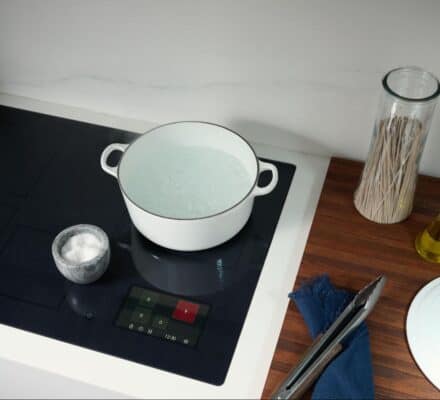 A white pot with boiling water sits on a black Wolf induction cooktop next to a small bowl of salt, a glass jar of spaghetti, olive oil, and a white pot lid on a wooden counter.