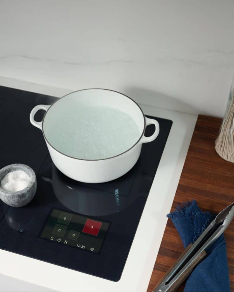 A white pot with boiling water sits on a black Wolf induction cooktop next to a small bowl of salt, a glass jar of spaghetti, olive oil, and a white pot lid on a wooden counter.