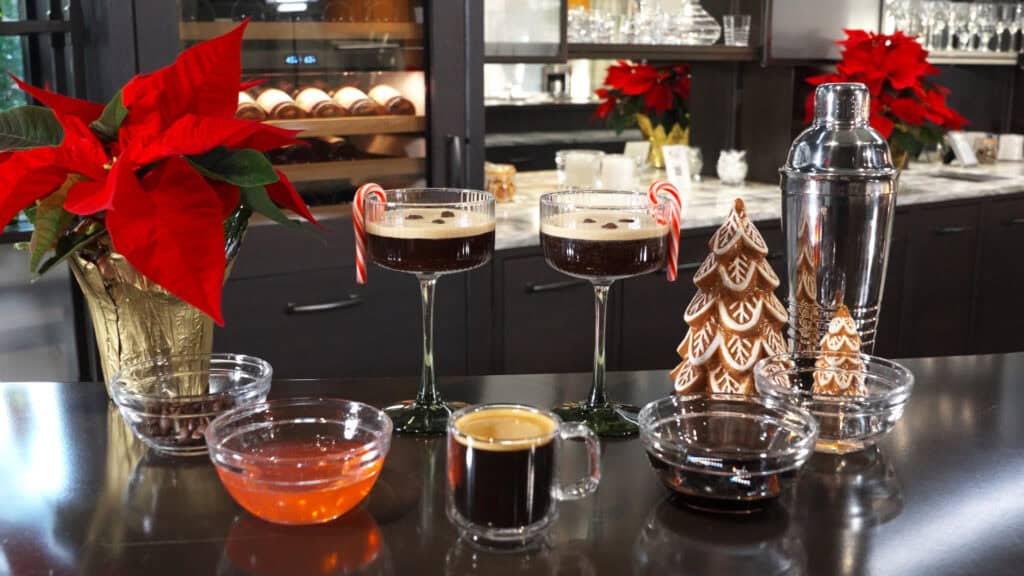 Two glasses of Peppermint Espresso Martini with candy canes, surrounded by ingredients, gingerbread trees, poinsettias, and a cocktail shaker on a kitchen counter.