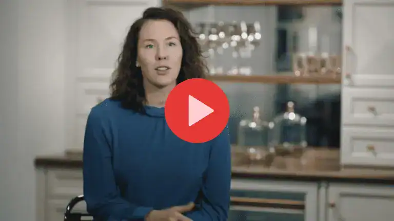 A woman with brown hair wearing a blue top sits in a Clarke kitchen, speaking; several glasses and jars are visible on shelves behind her.
