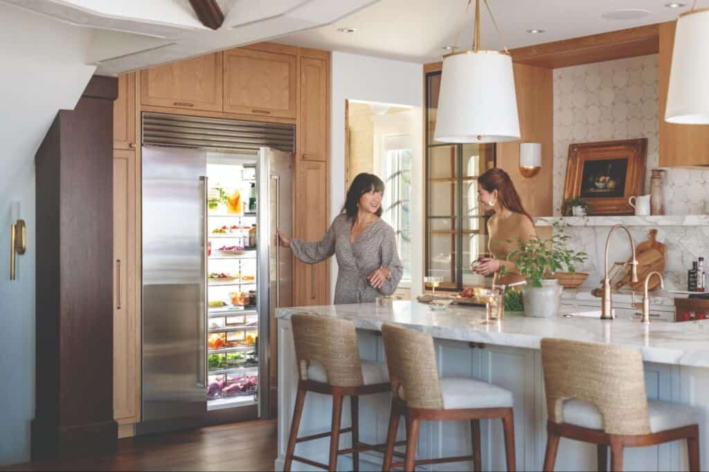 Two women stand in a modern kitchen showroom; one opens a built-in refrigerator while the other prepares food at a marble island countertop.