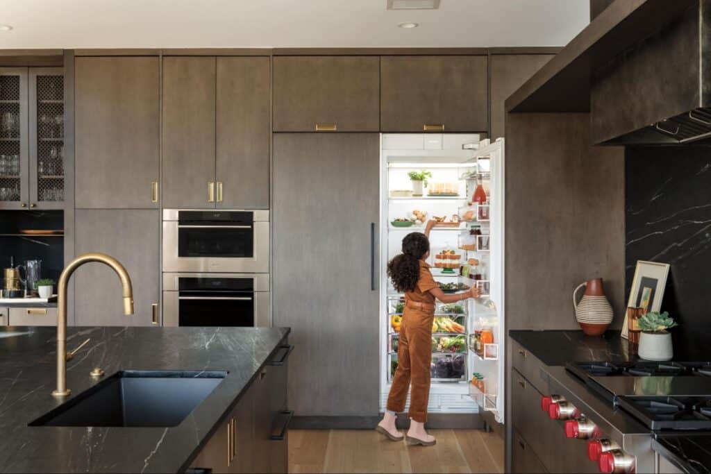 A person stands in a kitchen showroom with dark cabinets and marble countertops, looking inside an open refrigerator filled with food.