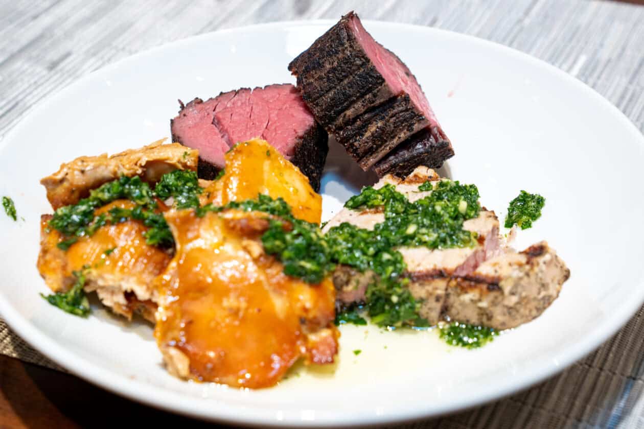 A white plate with slices of grilled steak, roasted potatoes, and grilled chicken topped with green herb sauce on a striped placemat—perfect examples of Wolf Product Demo Entrees and Sides.