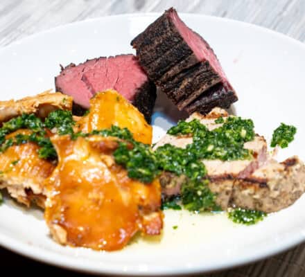 A white plate with slices of grilled steak, roasted potatoes, and grilled chicken topped with green herb sauce on a striped placemat—perfect examples of Wolf Product Demo Entrees and Sides.