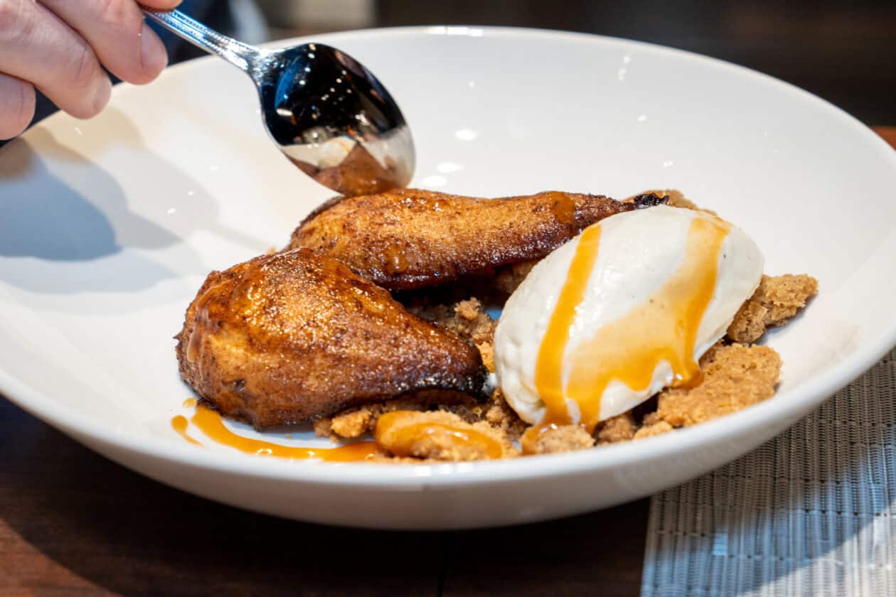 A person uses a spoon to serve a dessert with baked pears, vanilla ice cream, crumbled topping, and caramel sauce on a white plate—an elegant finish to any Wolf Product Demo Entrees and Sides experience.