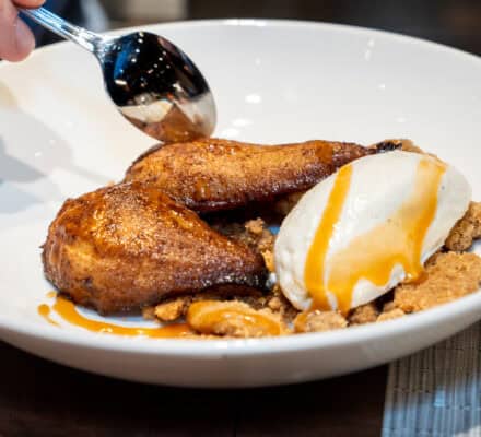 A person uses a spoon to serve a dessert with baked pears, vanilla ice cream, crumbled topping, and caramel sauce on a white plate—an elegant finish to any Wolf Product Demo Entrees and Sides experience.