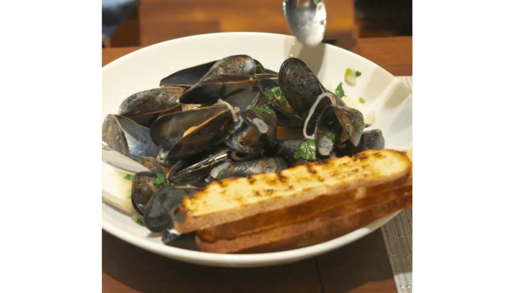 A bowl of steamed mussels in white wine, garnished with herbs and onions, served with two slices of grilled artisanal bread on a white plate.