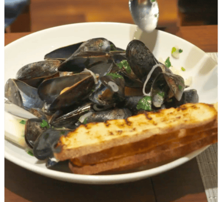 A bowl of steamed mussels in white wine, garnished with herbs and onions, served with two slices of grilled artisanal bread on a white plate.