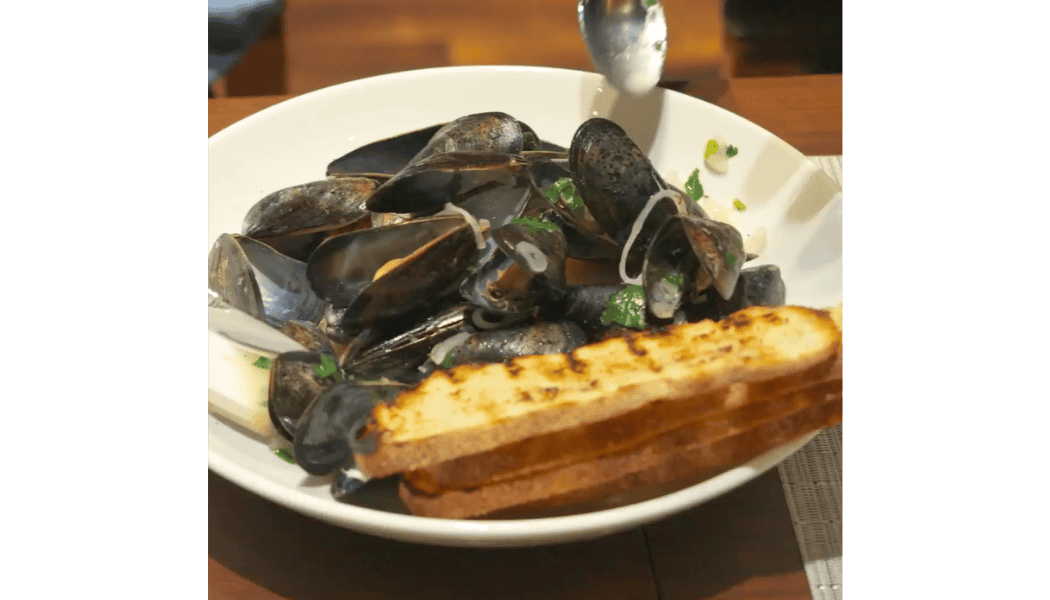 A bowl of steamed mussels in white wine, garnished with herbs and onions, served with two slices of grilled artisanal bread on a white plate.