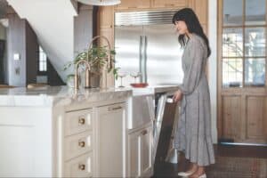 A woman in a long dress opens a dishwasher in a modern kitchen with marble countertops, wooden cabinets, and premium Wolf luxury kitchen appliances.