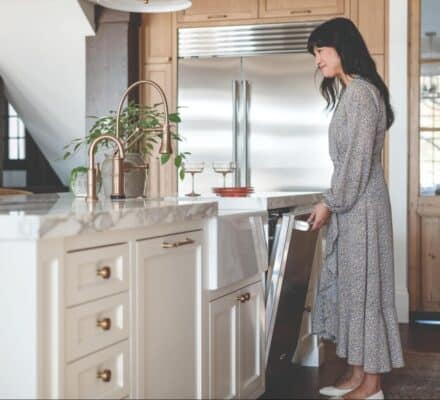 A woman in a long dress opens a dishwasher in a modern kitchen with marble countertops, wooden cabinets, and premium Wolf luxury kitchen appliances.