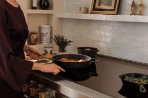 A person in a dark outfit cooks food in a pot on a modern Wolf stovetop, with vegetables in another pan and Sub-Zero kitchen appliances and other items elegantly displayed in the background.